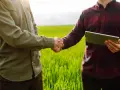 Dwóch mężczyzn ściska sobie dłonie na tle zielonego pola. Jeden z nich trzyma tablet, planując przyszłe zagospodarowanie przestrzenne.